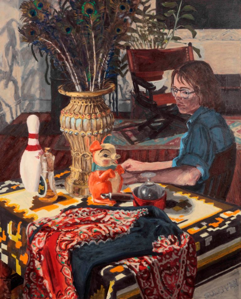 A middle-aged man wearing a blue shirt and shaggy brown hair sits in his studio at a table with a bowling pin, piggy bank, vase of peacock feathers, and a patterned table cloth.